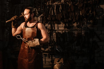 Portrait of a brutal muscular blacksmith standing in a dark workshop holding a vise and a hammer