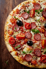 Pizza with sausage, cherry tomatoes, black olives and herbs. Home made food. Concept for a tasty and hearty meal. Brown wooden background. Top view.