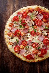 Pizza with ham, cheese, cherry tomatoes, champignon mushrooms and fresh herbs. Brown wooden background. Top view. 