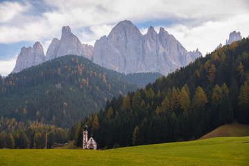 Fototapeta na wymiar Typical scenery of small Church of San Giovanni in Ranui with dolomites Odle in background and clouds and foliage