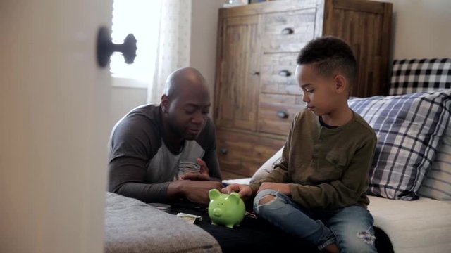 Father and son depositing coins into piggy bank in bedroom