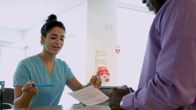 Female Nurse Explaining Paperwork To Male Patient At Clinic Reception