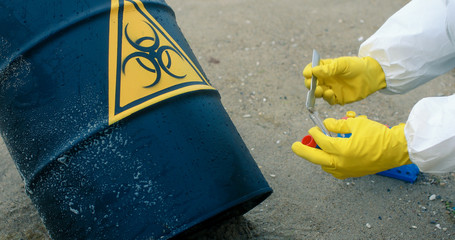 Close-up of a scientist`s hands in yellow protective gloves taking a sand sample with a spatula next to a barrel of bio-hazardous substances.