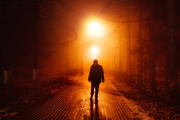 Sad man alone walking along the alley in night foggy park. Front view