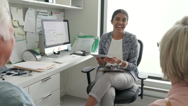 Happy Female Doctor With Digital Tablet Talking With Senior Couple Holding Hands In Clinic Doctors Office