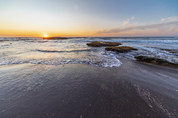 An amazing view of the sunset over the water in the Chilean coast. An idyllic beach scenery with the sunlight illuminating the green algae and rocks with orange tones and the sea in the background
