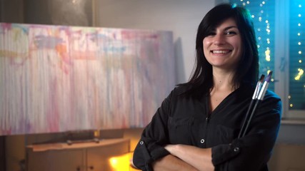 Portrait of amazing smiling female artist posing on illuminated background in art studio, large emotional painted picture on canvas staying on wooden easel, colorful vertical and horizontal strokes in - Powered by Adobe