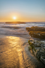 An amazing view of the sunset over the water in the Chilean coast. An idyllic beach scenery with the sunlight illuminating the green algae and rocks with orange tones and the sea in the background
