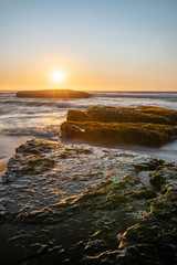 An amazing view of the sunset over the water in the Chilean coast. An idyllic beach scenery with the sunlight illuminating the green algae and rocks with orange tones and the sea in the background
