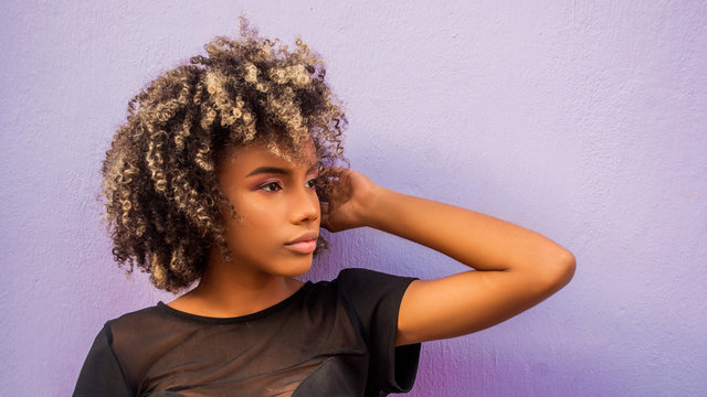 Mujer joven afrodecendiente cabello risado suelto con su braso levantado mostrando levemente su mano, mirando hacia un lado sobre fondo morado