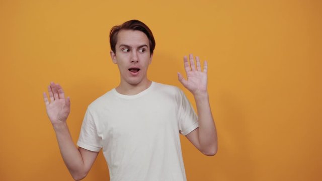 A man in a white t-short on yellow background gestures her hends with open mouth