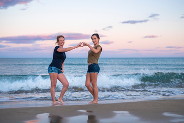 leisure and friendship concept - two happy smiling teenage girls or best friends at seaside making hand heart gesture at sunset