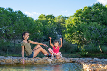 Beautiful mother and her little daughter outdoors. Beauty Mum and her Child playing with water in Park together at sunset. Outdoor Portrait of happy family. Mother's Day