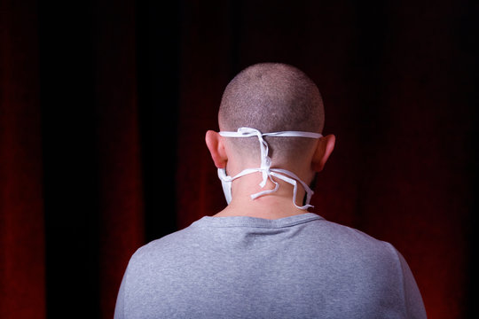 A Man In Medical Gauze Mask Standing Back To Camera On Dark Red Background. Virus Preventive Methods. Protection Of Upper And Lower Respiratory System From  Chinese Coronavirus Concept.