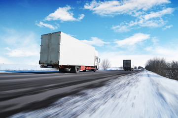 Two box trucks on the winter road in motion against a blue sky with clouds