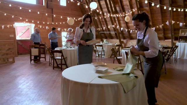 Female caterers setting tables, preparing for wedding reception in barn