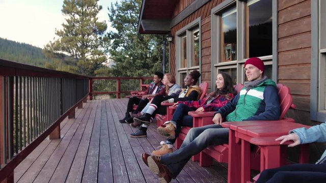 Young Friends Relaxing In Adirondack Chairs On Cabin Balcony