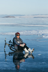 Fototapeta na wymiar Woman sitting near her bike on ice. The girl cyclist stopped to rest. She sits on the wheel and enjoys the beautiful view of the sunset. The traveler is ride a cycle. Female is riding bike on the ice.