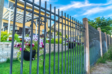 Black metal fence with potted colorful flowers against blurry homes and blue sky