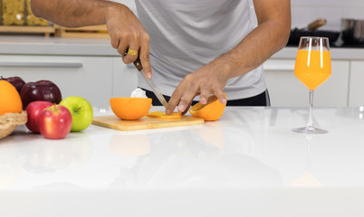 Young man sliced oranges on the wood cutting board in the kitchen, with orange juice in a beautiful glass, placed on the table as well.