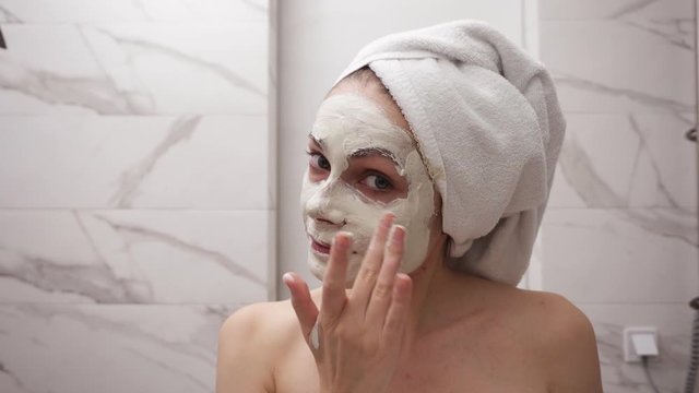 Portrait of young pretty woman with white towel on head is applying fresh white thick mask and scrub on the face. Natural organic cosmetics for skin care. Woman after shower, face skin care