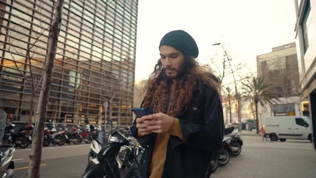 Portrait of long haired hipster walking on street in modern city in slow motion. He wears black jacket with leopard hoodie. He is typing on a cell phone.