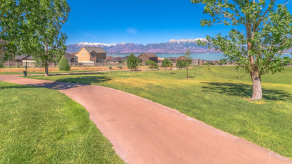 Obraz premium Panorama frame Narrow pathway on vast grassy field with lush trees and overlooking lake view