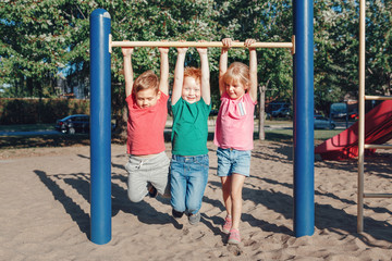 Fototapeta na wymiar Three funny Caucasian friends hanging on pull-up bars in park on playground. Summer outdoors activity for kids. Active children boys and girl doing exercises sport. Healthy happy childhood.