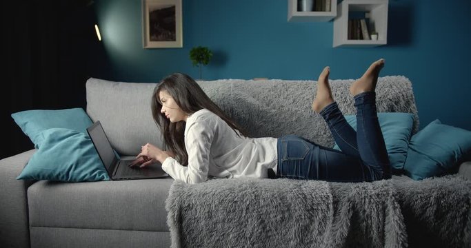 Full length portrait of stunning young girl with long dark hair in casual clothing lying on grey couch and surfing internet by using her laptop. Concept of leisure time and weekends at home