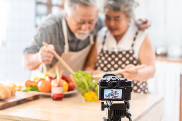 Couple senior Asian elder happy living in home kitchen. Grandfather eating salad dish after cook by grandmother recording vlog vdo for social blogger. Focus on camera. Modern lifestyle & relationship.