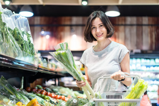 Young Asian beautiful woman holding grocery basket walking in supermarket. She is choosing green fresh vegetable picking up from shelf. Seen from side while she looking at products. Shopping concept.