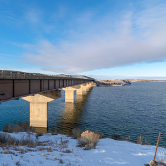 Obraz premium Square frame Bridge over a vast calm lake with snow covered shore during winter season