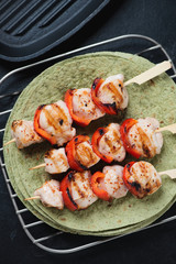 Wooden skewers with roasted chicken fillet and bell pepper slices on tortilla flatbread, vertical shot, above view