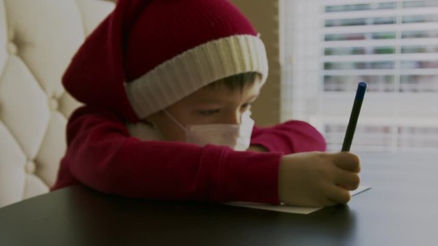 Sick little boy in a red sweater with red santa hat with the white pompon and in a white respirator mask on his face sitting at a table and holding a pen in his hands and writing or drawing on a blank