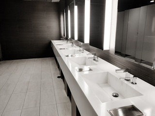 Public empty restroom with washstands, baby changer, and toillets in mirror. White sink row with...