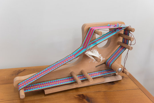 A small, tabletop, inkle loom on which a band, or strap, is in the process of being woven. - Powered by Adobe