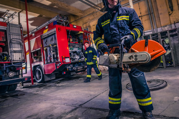 Fototapeta premium A fireman with uniform and helmet holding a chainsaw with fire truck in the background
