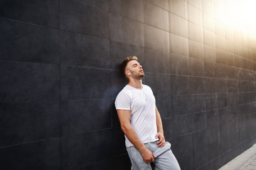 Attractive fit caucasian sportsman in tracksuit and in t-shirt leaning on the wall and resting. In ears are earphones.