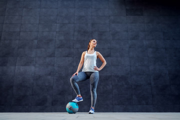 Fototapeta premium Attractive fit Caucasian brunette in sportswear holding foot on weight ball and posing outdoors. In background is dark wall.