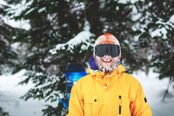 Fototapeta na wymiar Close up portrait of bearded snowboarder or skier wearing black goggles and orange hat with snow on face standing in mountain forest and smiling. Happy people vacation.