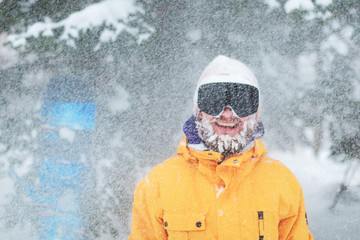 Fototapeta na wymiar Skier's or snowboarder portrait. Bearded man in winter hat and black goggles at snow blizzard in the forest. Happy and smiling snowboarder with snow on his face.