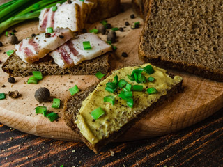 Slices of salted lard on rye bread with mustard and green onions spread on a cutting board on the surface of a wooden table with black pepper.