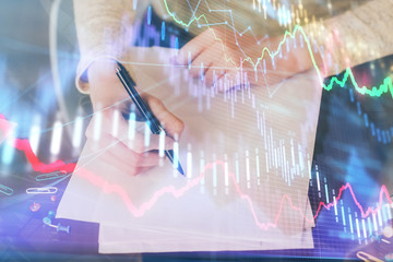 Hand taking notes in notepad. Forex chart holograms in front. Concept of research. Multi exposure