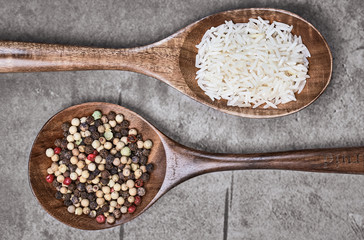 Mixed peppercorns and uncooked rice on two brown and wooden spoons lying on a gray textured surface. Top view and macro food photography.