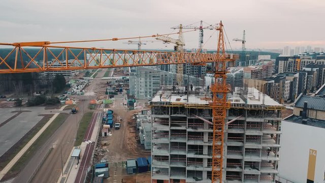 Slab-constructed apartment blocks, city-houses and town-houses building by workers. Aerial view from drone flying near crane on construction site against backdrop of modern urban area.