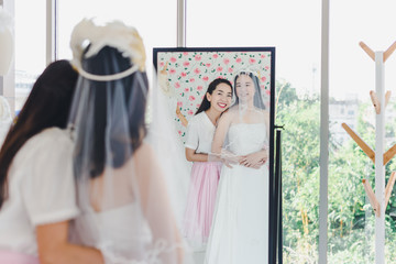 The bridesmaid is helping the beautiful bride wearing a white wedding dress in front of the mirror.