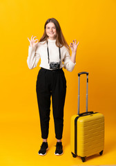 Full body of traveler teenager girl with suitcase over isolated yellow background showing an ok sign with fingers