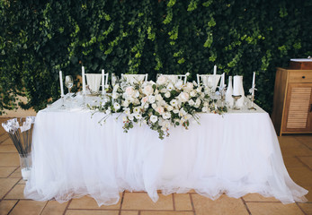 A wedding table with a white tablecloth, candles, a large bouquet of white roses and vintage chairs stands against a background of green foliage of wild grapes. Outdoor, summer ceremony.