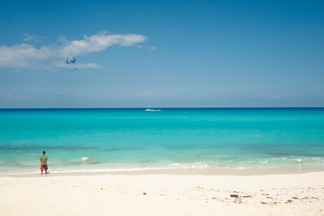 The turquoise sea and the white sand of the dolphin beach in Cancun Mexico, a man observes the horizon, in the background a parasailing with tourists in the blue sky