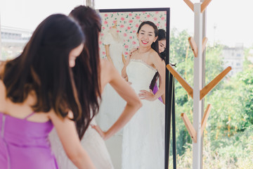 The bridesmaid is helping the beautiful bride wearing a white wedding dress in front of the mirror.